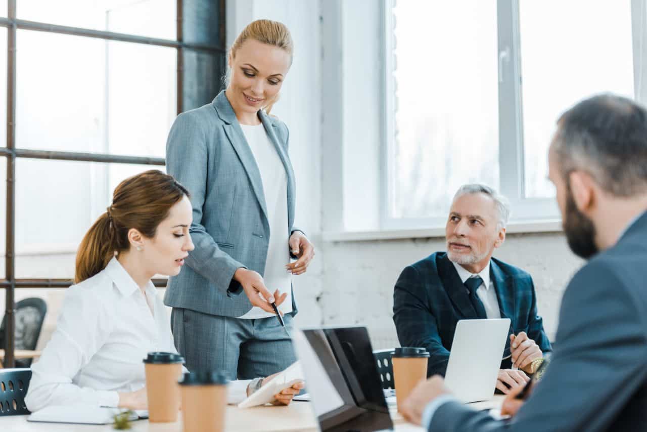 cheerful business coach standing near coworkers and gesturing near digital tablet