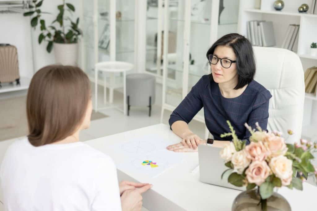 two young women conduct a coaching session in home office in light interior with laptop and flowers